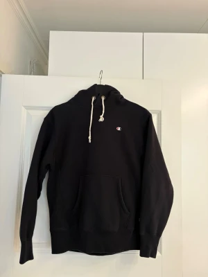 Svart hoodie från Champion - Svart hoodie från Champion med klassisk logga på bröstet och ärmen. Hoodien har vit dragsko i huvan och en stor magficka framtill. Tillverkad i mjukt bomullsmaterial och har ribbade muddar vid ärmslut och nederkant. 