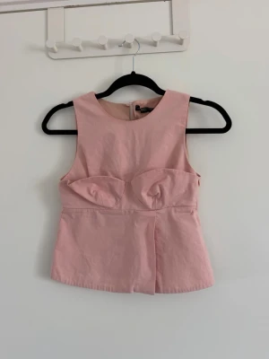 Ljusrosa ärmlös topp med kupdetalj - Snygg ljusrosa ärmlös topp med markerade kupor och rund halsringning. Toppen har en figurnära passform och dragkedja i ryggen. Perfekt för dig som vill ha en trendig och feminin look. Sparsamt använd. Är fläckig vid urringningen men inget som syns jätte tydligt