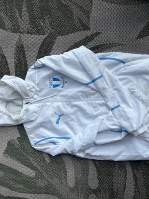 Vit vindjacka Malmö FF x Puma - Vit vindjacka med huva från Puma i samarbete med Malmö FF. Jackan har ljusblå detaljer och loggor på bröstet samt dragkedja framtill. Perfekt för blåsiga dagar och sportiga outfits.