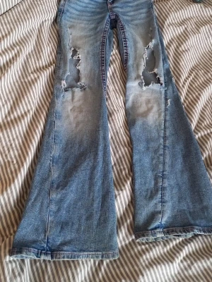 Blå slitna bootcut jeans - Säljer ett par blå bootcut jeans med slitningar och hål på båda benen framtill. Jeansen har en ljus tvätt och broderade detaljer på bakfickorna. Perfekta för dig som gillar en avslappnad och trendig stil.