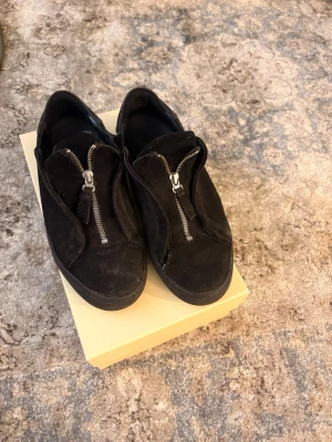 Svarta sneakers med dragkedja - Säljer ett par svarta sneakers i mocka med coola dragkedjor framtill istället för klassiska snören. Skorna har en låg profil och en chunky sula som ger en modern vibe. Perfekta för dig som gillar stilrena men ändå unika detaljer.