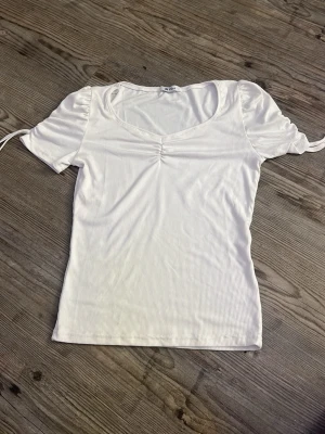 Vit ribbad topp med snörning - Säljer en vit ribbad topp med korta puffärmar och snörning i ärmsluten. Toppen har v-ringning och en liten rynkdetalj framtill. Perfekt till jeans eller kjol för en fräsch look.
