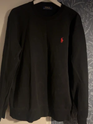 Svart sweatshirt från Polo Ralph Lauren - Klassisk svart sweatshirt från Polo Ralph Lauren med rund halsringning och den ikoniska röda logotypen broderad på bröstet. Tröjan har långa ärmar och ribbade muddar vid ärmslut och nederkant. Perfekt för en clean och stilren look.