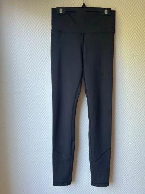 Svarta sporttights med hög midja - Säljer ett par svarta sporttights med hög midja och perforerade detaljer vid bensluten. Tightsen är tillverkade i ett stretchigt funktionsmaterial som passar perfekt för träning eller löpning. Snygg och enkel design.