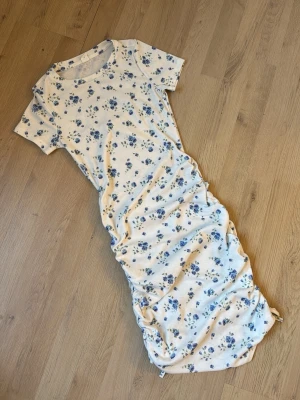 Blommig långklänning med snörning - Ljus långklänning med blå blommigt mönster och korta ärmar. Klänningen är kroppar men det finns också snören längst ner på klänningen där man kan justera hur man vill ha den!! Perfekt till sommaren 