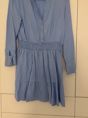 Ljusblå klänning med knappar - Supersöt ljusblå klänning med långa ärmar, v-ringning och knappar framtill. Klänningen har smock i midjan för en snygg passform och volang i kjolen. Perfekt för dig som gillar en avslappnad men ändå stilren look.