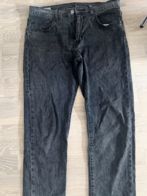 Svarta raka jeans från Levi's - Säljer ett par svarta raka jeans från Levi's 502  med klassisk femficksmodell och knappgylf. Jeansen har en diskret tvättad look och Levi's ikoniska läderpatch bak i midjan. Perfekta till en avslappnad streetwear-stil. Storlek 33/32