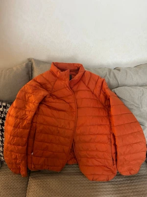 Orange pufferjacka med hög krage - Säljer en snygg orange pufferjacka med hög krage och quiltad design. Jackan har dragkedja framtill och är lätt vadderad för extra värme. Perfekt för kalla vinterdagar och ger en cool look till din outfit.