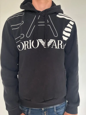Svart hoodie från Emporio Armani - Svart hoodie med stor vit EA7-logga och grafiska detaljer framtill. Huva och känguruficka. Snygg streetstil som passar perfekt till jeans eller joggers. Materialet är mjukt och bekvämt, perfekt för chill dagar.