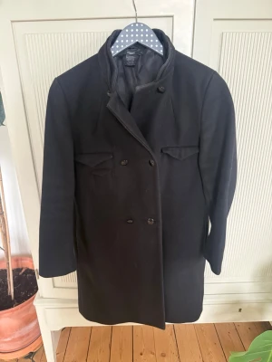 Svart dubbelknäppt ullkappa Flippa K - strl xs - Stilren svart kappa i ullblandning med dubbelknäppning och två fickor med lock framtill. Klassisk rak passform och hög krage som ger en tidlös look. Perfekt för kyliga dagar och enkel att matcha med olika outfits. Slitage i foder vid fickor. Förövrigt bra skick. 