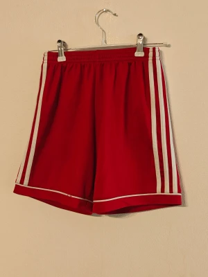 Adidas träningsshorts - Snygga röda shorts från Adidas med klassiska vita ränder längs sidorna. Tillverkade i ett lätt och bekvämt syntetmaterial som andas, perfekta för sport eller chill. Elastisk midja för skön passform och sportig look.