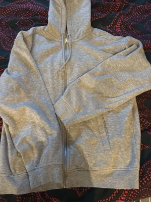 Ljusgrå zip hoodie med huva - Säljer en ljusgrå hoodie med dragkedja och huva. Tröjan har två fickor framtill och ribbade muddar vid ärmslut och nederkant. Perfekt för chill dagar eller när du vill ha något bekvämt och enkelt.