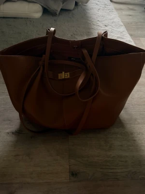 Väska Gina Tricot - Snygg och rymlig shoppingväska från Gina Tricot i brunt konstläder. Väskan har dubbla handtag, dekorativt spänne framtill och innerfack med dragkedja. Perfekt för dig som vill ha plats för allt du behöver under dagen.