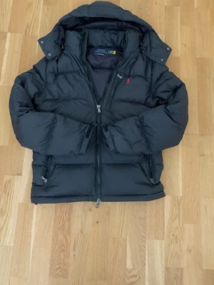 Svart pufferjacka med huva - Snygg svart pufferjacka från Polo Ralph Lauren med huva och dragkedja framtill. Jackan har två sidofickor med dragkedja och den klassiska röda loggan på bröstet. Perfekt för kalla vinterdagar tack vare det tjocka, vadderade materialet. Priset går att diskutera vid snabb affär 