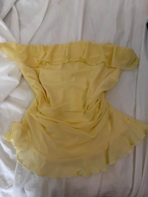 Gul offshoulder volangklänning - Söt gul axelbandslös korttröja med volangdetaljer upptill och nedtill. Tröjan har en luftig och lätt känsla, perfekt för varma dagar. Materialet ser ut att vara polyester eller liknande syntet. Passformen är tight och modellen är offshoulder för en trendig look.