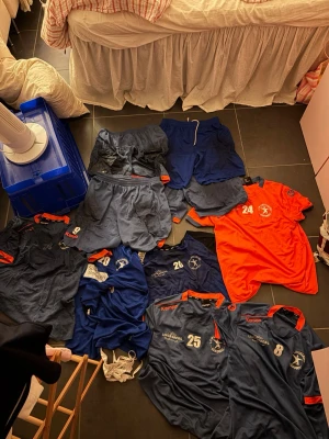 Handbollskläder i blått och orange - Säljer ett set med handbollskläder i marinblått och orange. Inkluderar flera matchtröjor med nummer och logga, shorts och en orange träningströja. Kläderna har tryckta detaljer och är tillverkade i funktionsmaterial som andas. Perfekt för träning eller match.