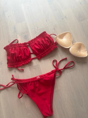 Röd bikini med volang och snörning XS - Snygg röd bikini i bandeau-modell med volangdetaljer och snörning både på toppen och trosan. Uttagbara inlägg medföljer. Perfekt för sommaren och strandhäng!
