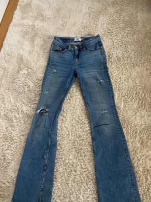 Blå bootcut jeans med slitningar - Säljer ett par blå bootcut jeans med snygga slitningar på benen och klassiska fem fickor. Jeansen har en något utsvängd passform nedtill och detaljer i orange söm. Mid waist