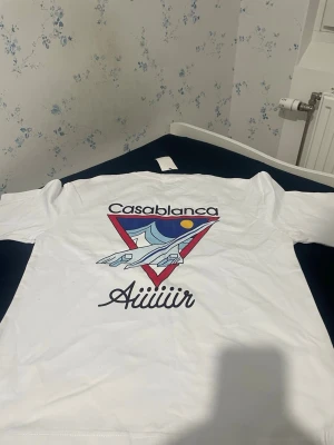 Vit Casablanca t-shirt med tryck - Säljer en vit t-shirt från Casablanca med stort färgglatt tryck på ryggen som visar ett flygplan och texten 'Casablanca Aiiiir'. Framtill finns en mindre textdetalj. Klassisk passform och korta ärmar, perfekt till sommaren. Vi kan komma överens med priset