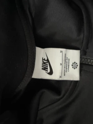 Svart Nike träningströja med dragkedja - Svart långärmad träningströja från Nike med halv dragkedja och ficka framtill. Snygga detaljer med Nike-loggor längs ärmarna i vitt och rött. Perfekt för träning eller chill. Materialet är mjukt och stretchigt för maximal komfort.