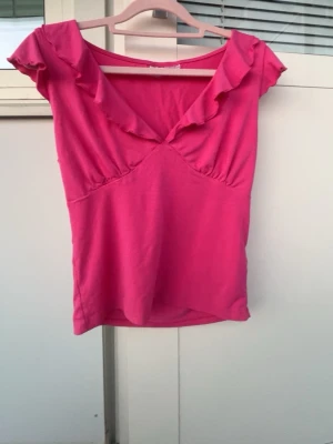 Rosa topp med volangdetalj - Snygg rosa topp med korta ärmar och volangdetalj längs den djupa v-ringningen. Toppen har en feminin passform och är tillverkad i mjukt bomullstyg. Perfekt för dig som vill ha en färgklick i garderoben.