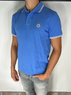 Moncler Piké  - Snygg blå pikétröja med vit kant på krage och ärmslut. Tröjan har korta ärmar, knappslå framtill och en liten logga på bröstet. Perfekt till jeans eller shorts för en clean och fräsch look.