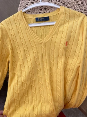 Gul kabelstickad tröja Polo Ralph Lauren - Gul kabelstickad tröja från Polo Ralph Lauren med V-ringning och klassisk broderad logga på bröstet. Tröjan har långa ärmar och ribbade muddar vid ärmslut och nederkant. Perfekt för dig som gillar preppy stil och vill ha något färgglatt i garderoben.