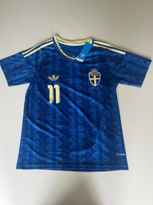 Blå Sverige fotbollströja Elanga 11 - Snygg blå fotbollströja från Adidas med Sveriges landslagsemblem och nummer 11 på bröstet. Elanga och nummer 11 står på ryggen. Tröjan har gula detaljer, korta ärmar och klassiska Adidas-ränder på axlarna. Perfekt för dig som vill stötta Sverige på match!