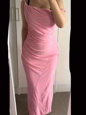 Lång rosa klänning med drapering - Säljer en lång rosa klänning med draperad effekt och smala axelband. Klänningen har en asymmetrisk axel och figurnära passform som framhäver siluetten. Perfekt för dig som vill ha en feminin och trendig look.