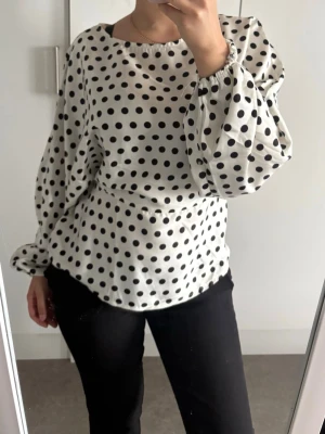 Prickig vit blus med knytband - Snygg vit blus med svarta prickar och ballongärmar. Blusen har rund halsringning, knytband i midjan och är tillverkad i ett glansigt, mjukt material som ger en lyxig känsla. Perfekt för dig som gillar trendiga detaljer och mönster.