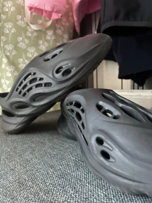 Yeezy  - Säljer ett par svarta tofflor i foam-material med futuristisk design och många hål för ventilation. De har en chunky sula och är perfekta för att glida runt hemma eller ute. Lätta och bekväma att bära, passar dig som gillar streetwear-vibe.