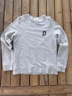 Grå sweatshirt från Dondup - Snygg grå sweatshirt från Dondup med rund halsringning och svart D-brodyr på bröstet. Tröjan har långa ärmar och ribbade muddar vid ärmslut och nederkant. Perfekt för en avslappnad och stilren look.