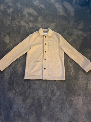 Beige overshirt  - Stilren beige overshirt från Bläck med tre stora utanpåliggande fickor framtill och knäppning med svarta knappar. Klassisk krage och raka ärmar. Perfekt lager-på-lager-plagg i mjukt tyg som ger en avslappnad look.