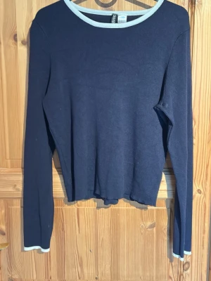 Mörkblå långärmad tröja med vit kant - En enkel och stilren mörkblå långärmad tröja med vita kanter vid halsringning och ärmslut. Tröjan har en rak passform och är tillverkad i ett mjukt, tunt material som passar perfekt till lager på lager. Snygg att matcha med jeans eller kjol. Det finns djur i huset så kan finnas spår av hundhår på tröjan som kan tvättas bort. 