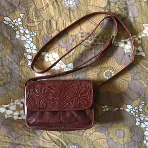 fin liten blomväska😽 - liten väska med ingraverat fint blommönster på framsidan. från indiska. stänges med magnetisk knapp och det finns en liten innerficka som stängs med dragkedja. bra använt skick:) axelremmen går att spänna till olika längder, det längsta är 122cm. mått: bredd: 16cm. höjd: 12cm. djup: 5cm. 