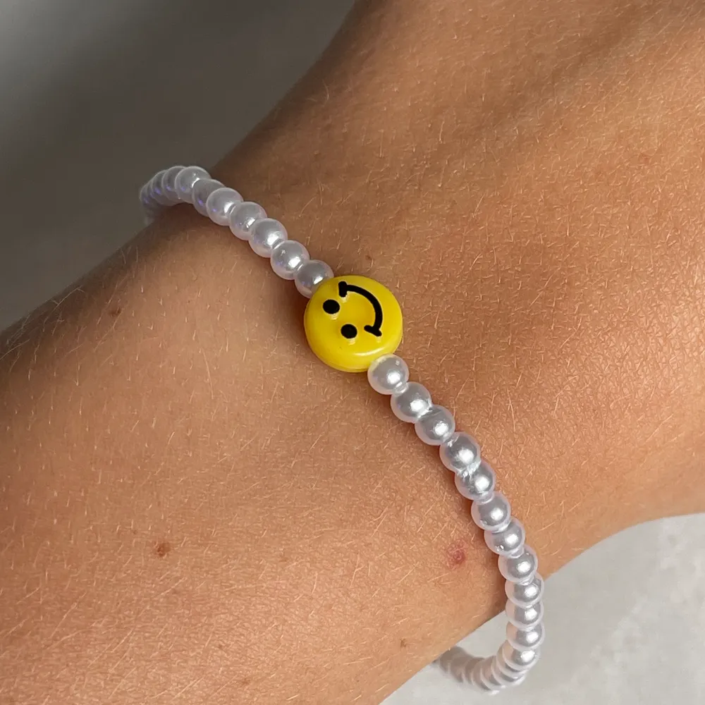 Ett jättefint och trendigt smiley armband med vita små pärlor och en smiley i mitten. Ingår spänne och ringödlor. Man väljer själv storlek. Accessoarer.