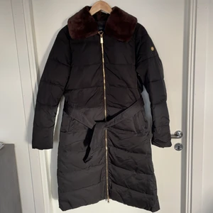 Jacka Morris str M  - Nästan helt ny Jacka från Moris. Fint kvalité och skick. Lång vadderad kappa från Morris Lady med knytbälte i midjan och avtagbar fuskpälskrage. Stängs med tvåvägs-metalldragkedja. Kappan har två snedställda fickor framtill som stängs med dolda tryckknappar, samt en innerficka.