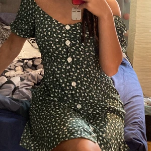 Grönblommig klänning - Kort grön klänning me vita blommor och knappar! Passar perfekt på en varm sommardag. Säljer då jag inte tyckte färgen passade min hudton, men det har väldigt par passform som visar kurvor och är lite lösare ner till. Man kan bära den som off-shoulder eller som vanligt.