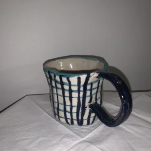 Handgjord vit kopp med blåa rutor - En handgjord kopp i keramik med ett vitt yttre men som är ofärgad inuti. Den har två nyanser av blått på ränderna samt samt ett blått öra. 