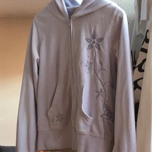 Hoodie - Snygg pastelllila hoodie med blomdetaljer. Den är i väldigt fint skick fortfarande väldigt mjuk i tyget. Skulle säga att den sitter bättre på folk som är korta(själv är jag 172 cm och den sitter precis vid armarna på mig) 