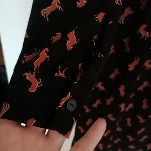 Klänning med volangkrage från Monki  - Helt rätt i höst - klänning med volangkrage, knäppning, vadlång, lagom vid och lagom figursmickrande. Svart med orangea hästmönster. Aldrig använd. Lätt puffärm och supertrendig. 