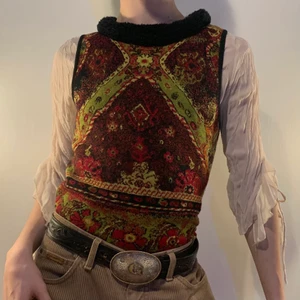 Röd grön svart orange stickat linne päls - Lev din höst dröm i detta stickade vintage linnet med det galnaste röda, gröna, orangea och svarta mönstret som påminner om en gammaldags matta 🎻  Kanten längst ner och vid axlarna är svarta vilket skär av mönstret på ett cleant sätt! Man får inte glömma den svarta otroliga fauxfurr kragen som gör detta linnet så lyxigt! Toppen är otroligt snygg till layering med en polo eller flowig blus under och är så så snygg till ett par bruna, röda eller gröna manchester byxor och ett statement bälte! 