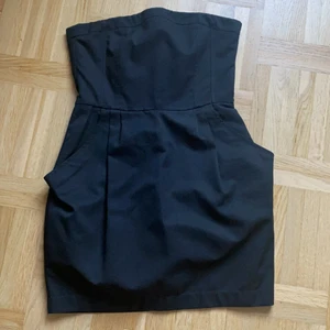 Den lilla svarta strl 38 - Snygg svart klänning i stlr 38. Axellös med en gummilinning överst i ringningen som ser till att klänningen inte åker ner. Två stora fickor framtill som snyggt går in i de sydda vecken.  