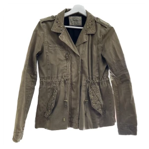 Zara - Armyjacka med nitar - Grön armyjacka med nitar från Zara. Strl M men liten modell, passar mig som S. Färgen är svårfångad, bild 2 fångar färgen bäst! Använt skick, se bild 3 där snöret är lite ”utdraget”. Slitna nitar. Bortklippt materiallapp.