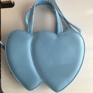 Ljusblå handväska - Söt handväska med hjärtan från kawaii.se. Oanvänd, fint skick. Avtagbar axelrem, rött foder samt liten innerficka. Dimensioner: 30cm bred, 20cm lång, 8cm djup. Priset inkluderar frakt, meddela vid frågor!