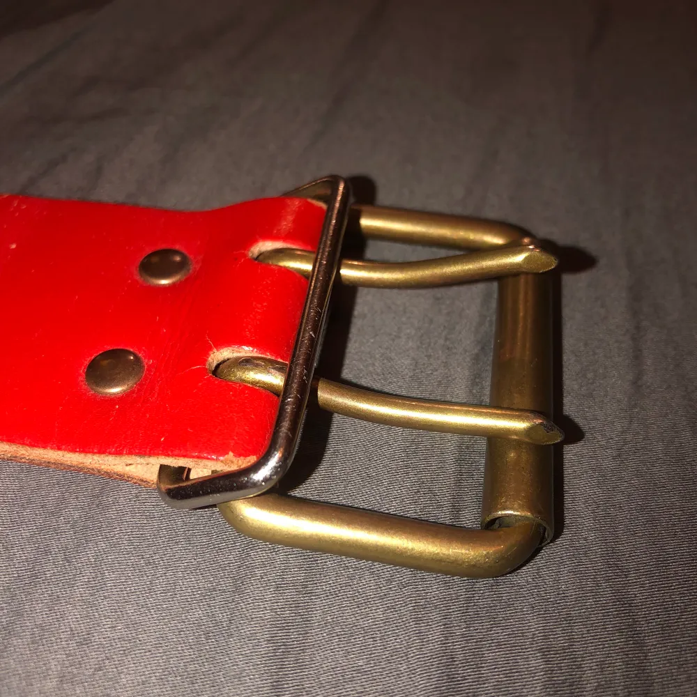 Rött läderaktigt skärp med dubbelknäppe i guldliknande metall. Längd 92cm.. Accessoarer.