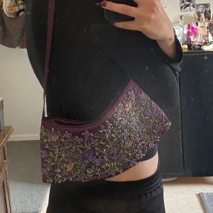 Lila väska - Säljer denn jättefina lila väska med broderade blommor på då den tyvärr inte kommer till användning :/ Superfin och med ett fack på insidan. Stängs med hjälp av en knapp. Köpt secondhand <3