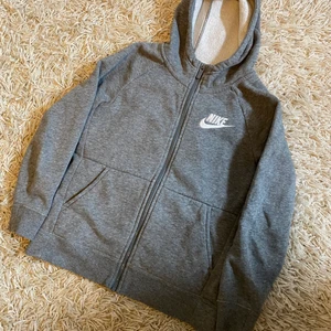Grå Nike hoodie med dragkedja  - Denna Nike hoodie är använd fåtal gånger, dragkedjan går väldigt lätt och är inte alls trög, är supersnygg med ett par tränings tights till och är i storlek 137-146/M 