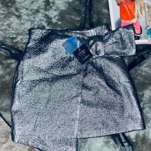 Kjol - Kort kjol silverstruktur fodrad inuti. Fakedetalj fram, går inte att justera. Stängs med dolt blixtlås bak. Aldrig använd.