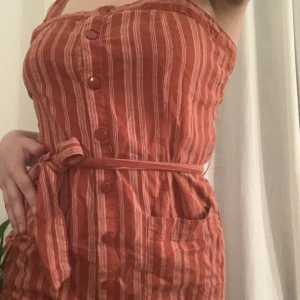 Klänning - Säljer denna supersöta klänning då den tyvärr  är för liten. Knappt använd. Röd-rosa randig. S/XS. Snöre/bälte runt magen och knappar hela vägen ner på klänningen sån går att öppna 